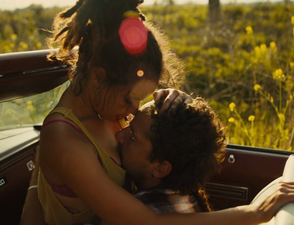 Two young people in a convertible share a tender moment, one resting their head on the other's chest as sunlight leaks softly into the lens, casting a golden warmth over the scene.
